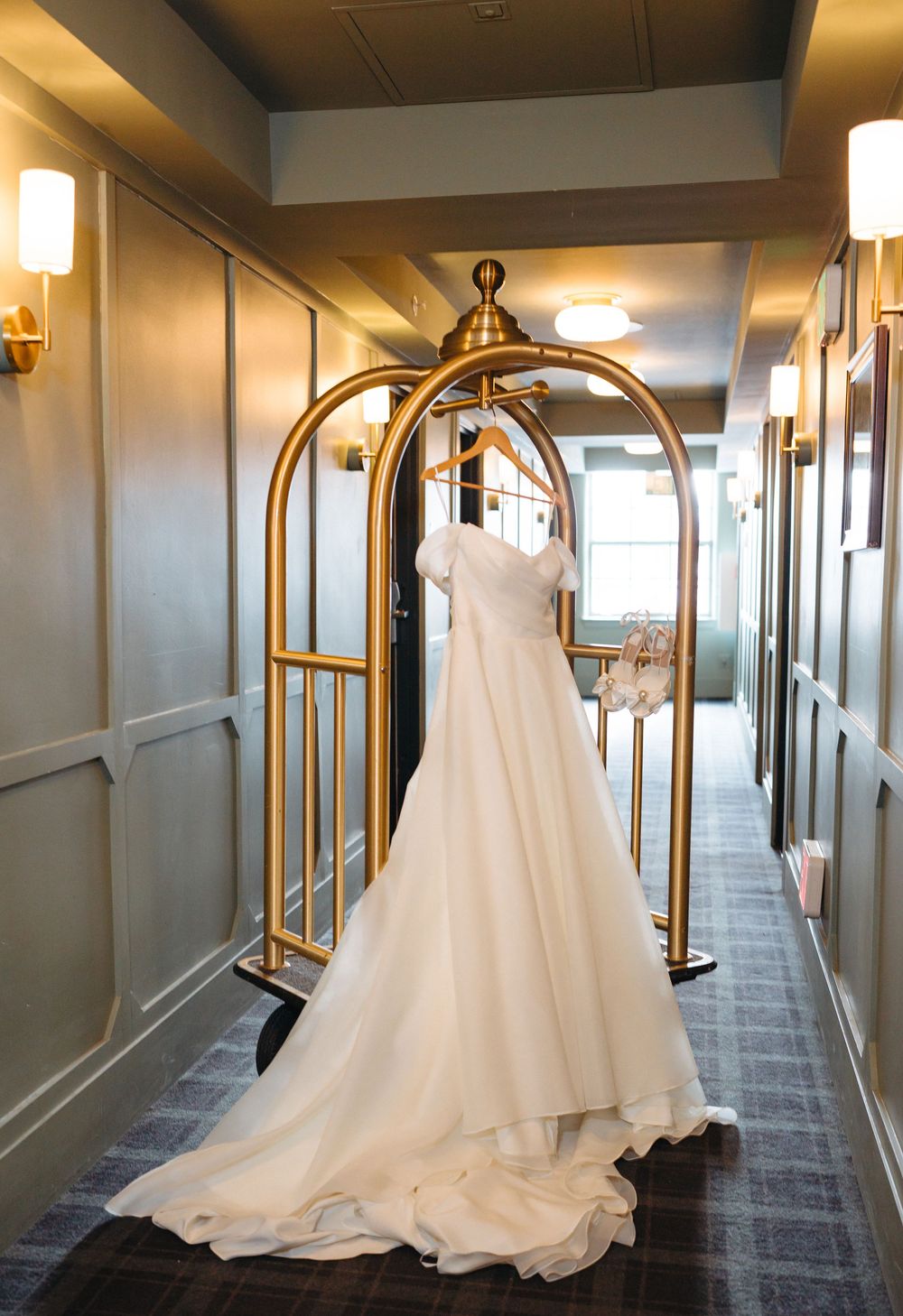 An elegant white wedding dress displayed on a gold luggage cart in a hotel hallway.