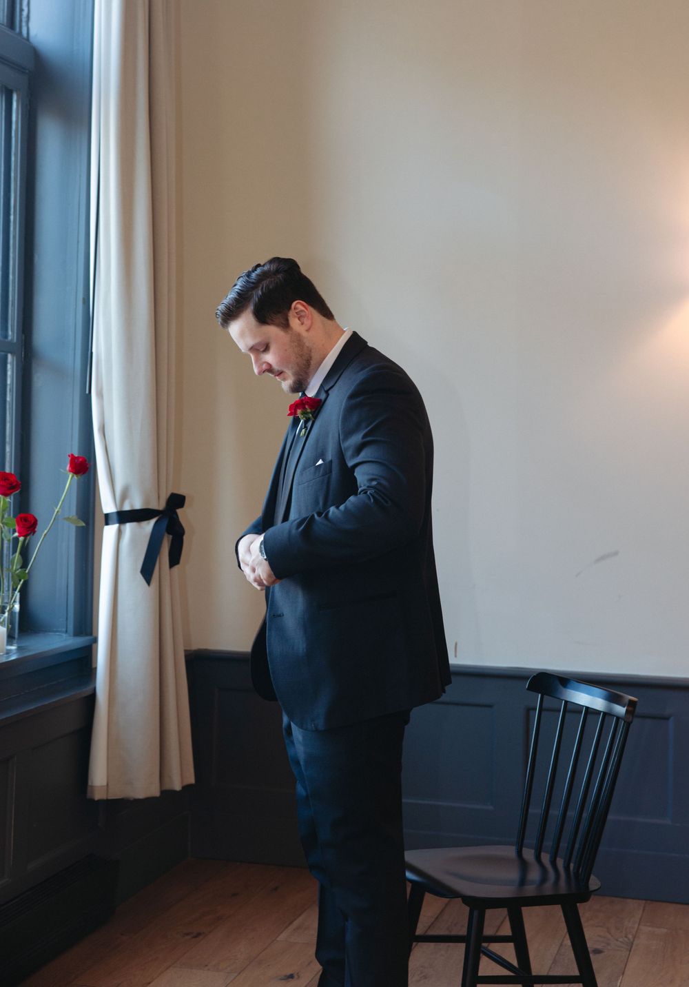 A person in a dark suit stands contemplatively near a wooden chair in a minimalist room with a single rose.