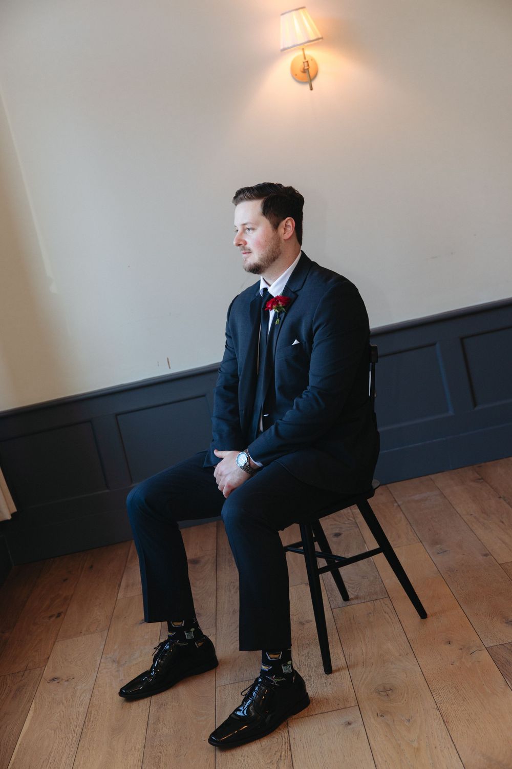 A person in a dark suit sits on a wooden stool against a wall with wainscoting and a sconce light.