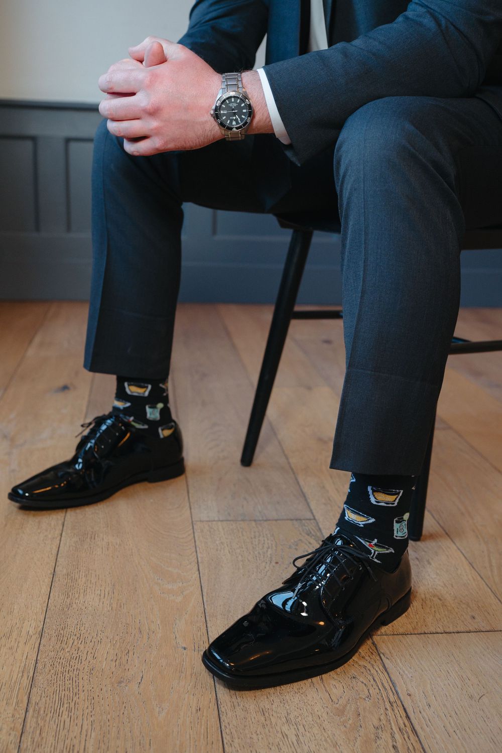 Close-up of black dress shoes, patterned socks, and a wristwatch on crossed legs against hardwood flooring.