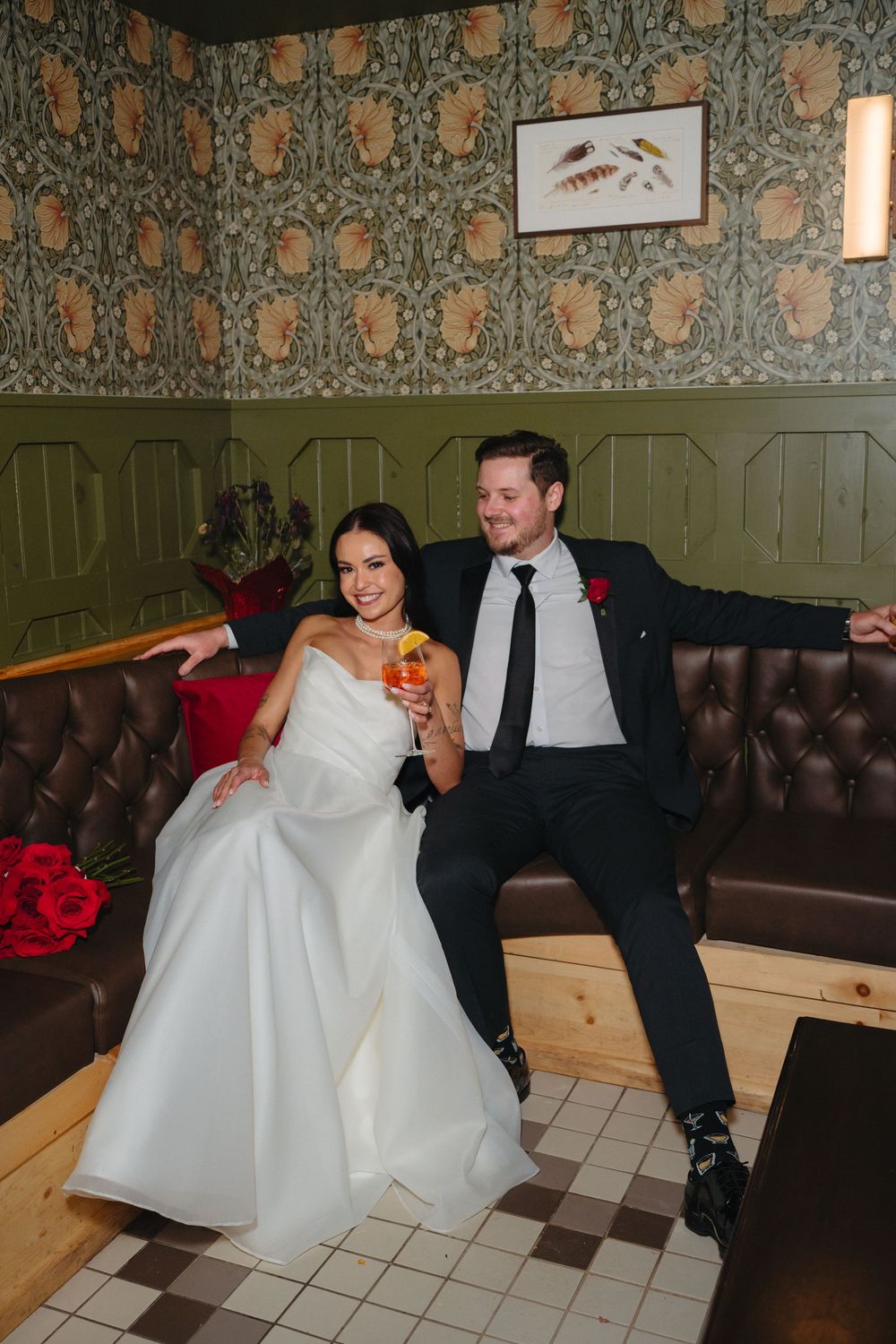 Wedding couple relaxes on leather booth seating with decorative wallpaper in restaurant setting.