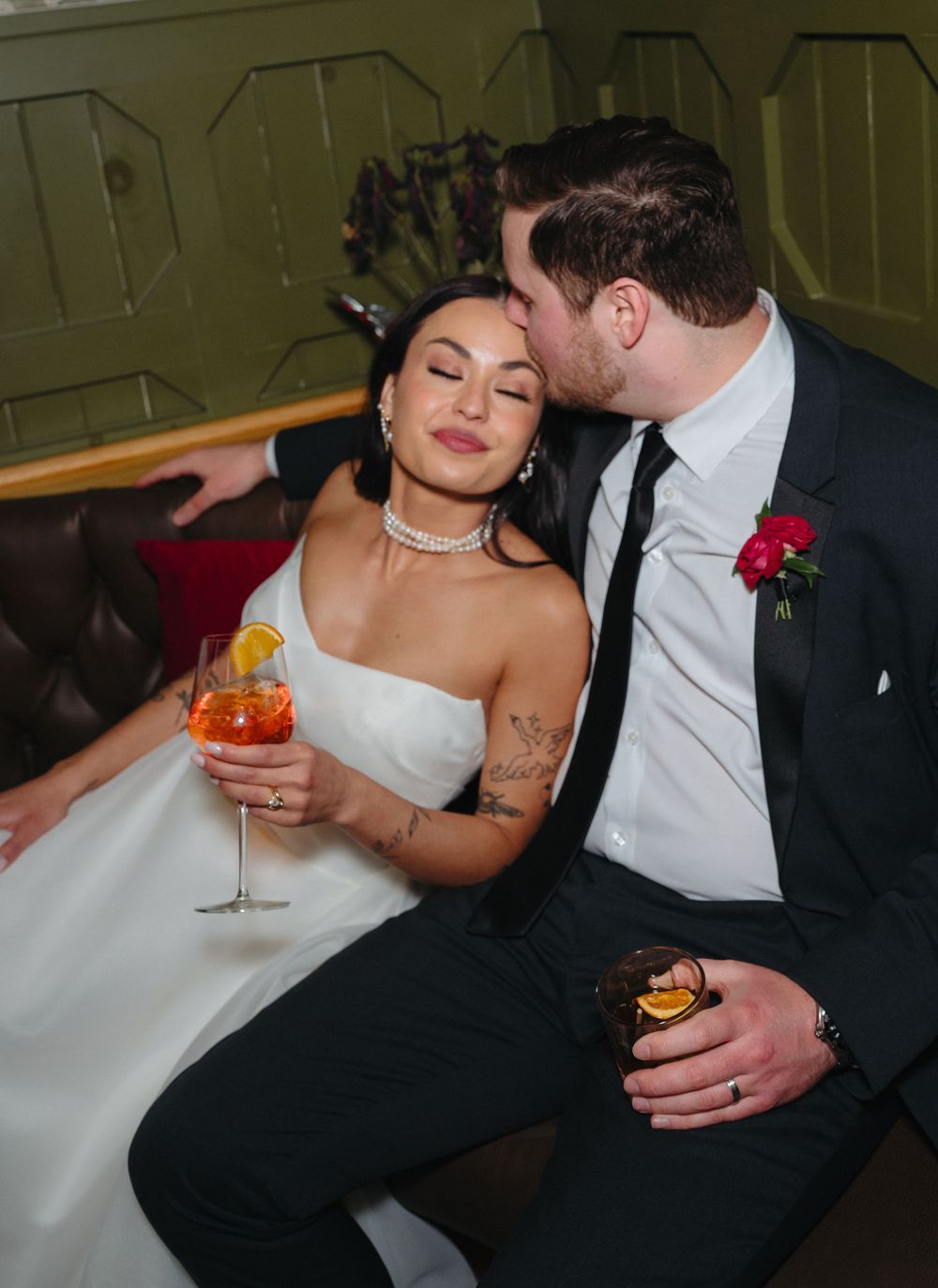 A wedding couple shares an intimate moment on a leather couch while holding cocktails.