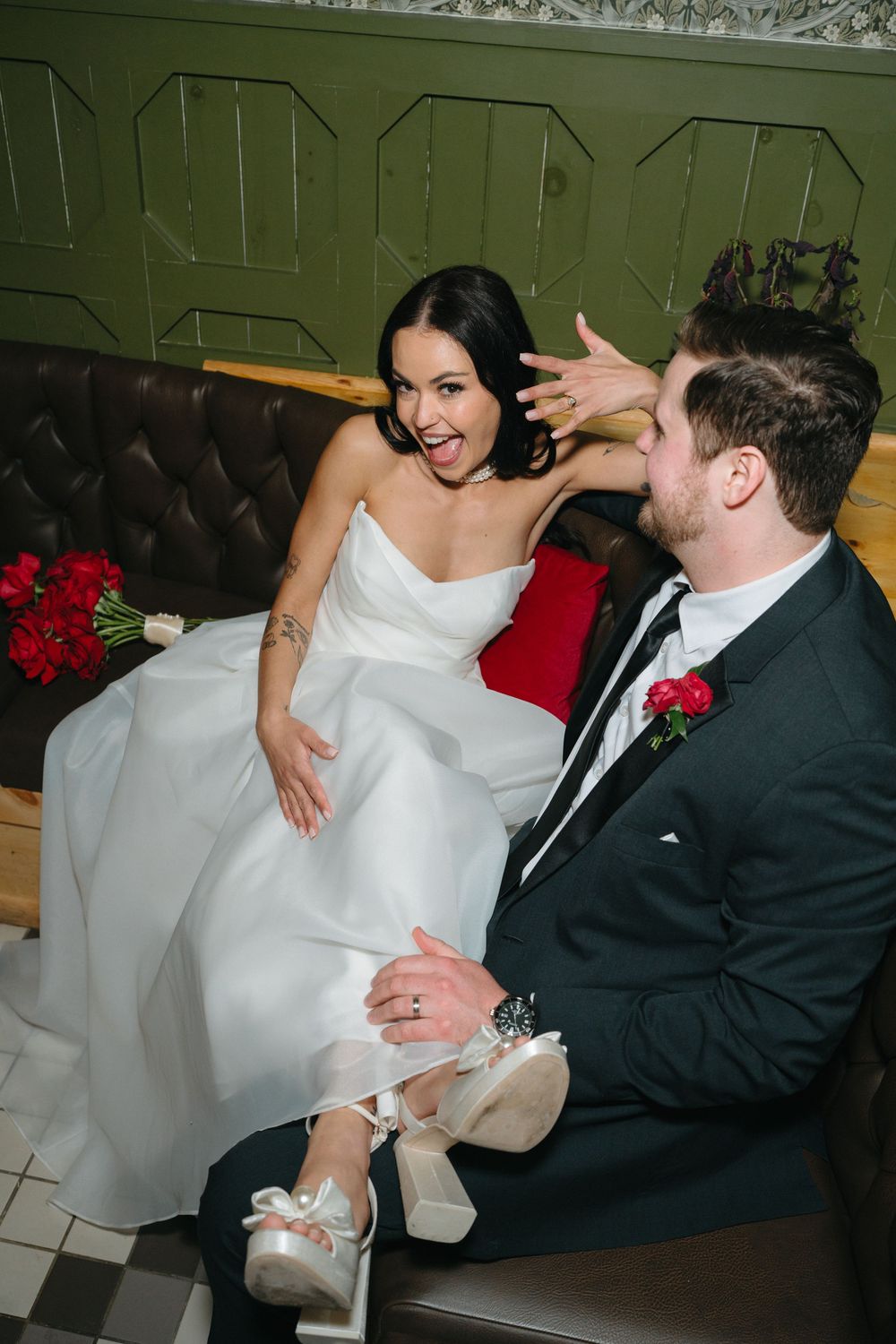 Newlyweds share a joyful moment on a green leather bench with red roses and elegant wedding attire.