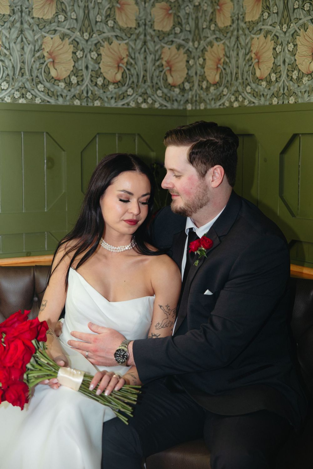 A romantic wedding couple shares an intimate moment on a bench with red roses against vintage wallpaper.