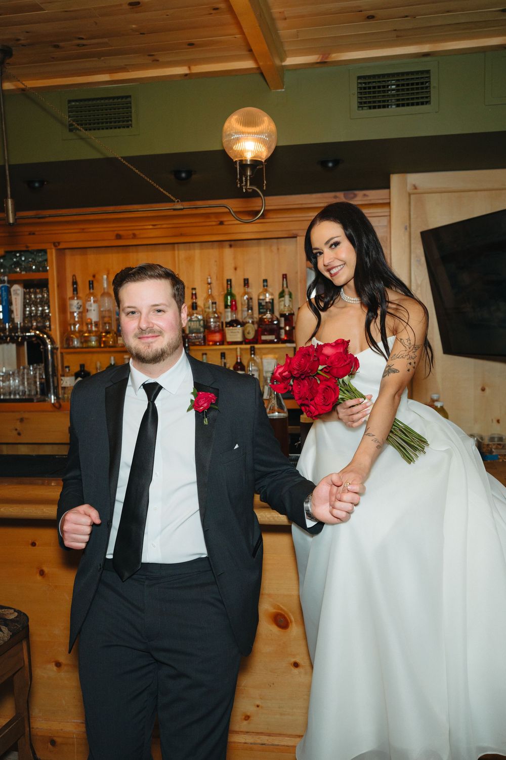 A wedding couple poses at a rustic wooden bar decorated with red roses and warm lighting.