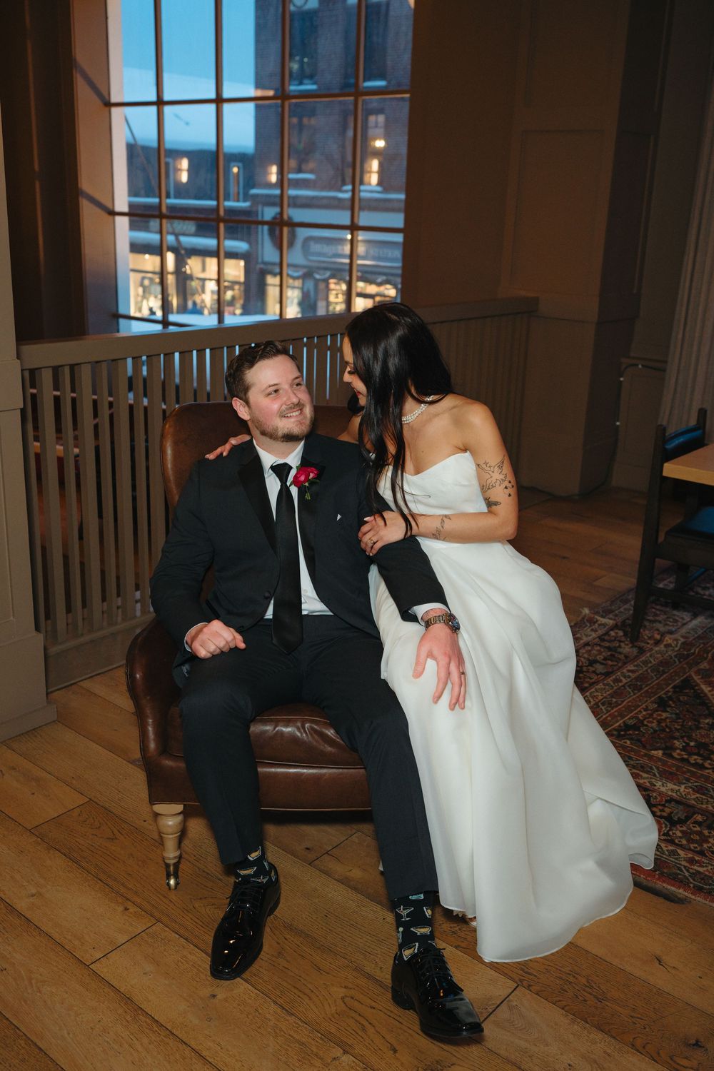 A romantic wedding moment with a couple sitting together on a vintage chair by a window at dusk.