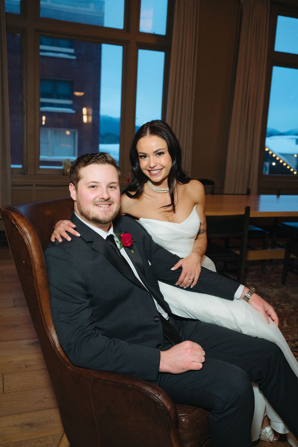 A newlywed couple relaxes together in a leather armchair by large windows during their wedding celebration.
