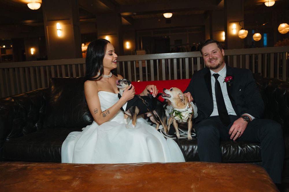 A bride in white dress and groom in dark suit sit together on a dimly lit bench at their wedding reception.