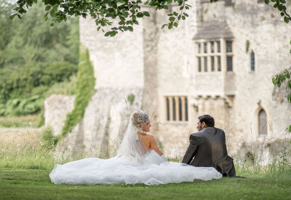 Bride and groom sit together on grass against ramparts of Allington castle during romantic wedding photo session.