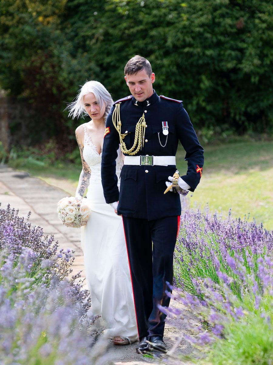 A couple in wedding attire walks through a beautiful garden path lined with blooming purple lavender flowers | Allington Castle in Kent