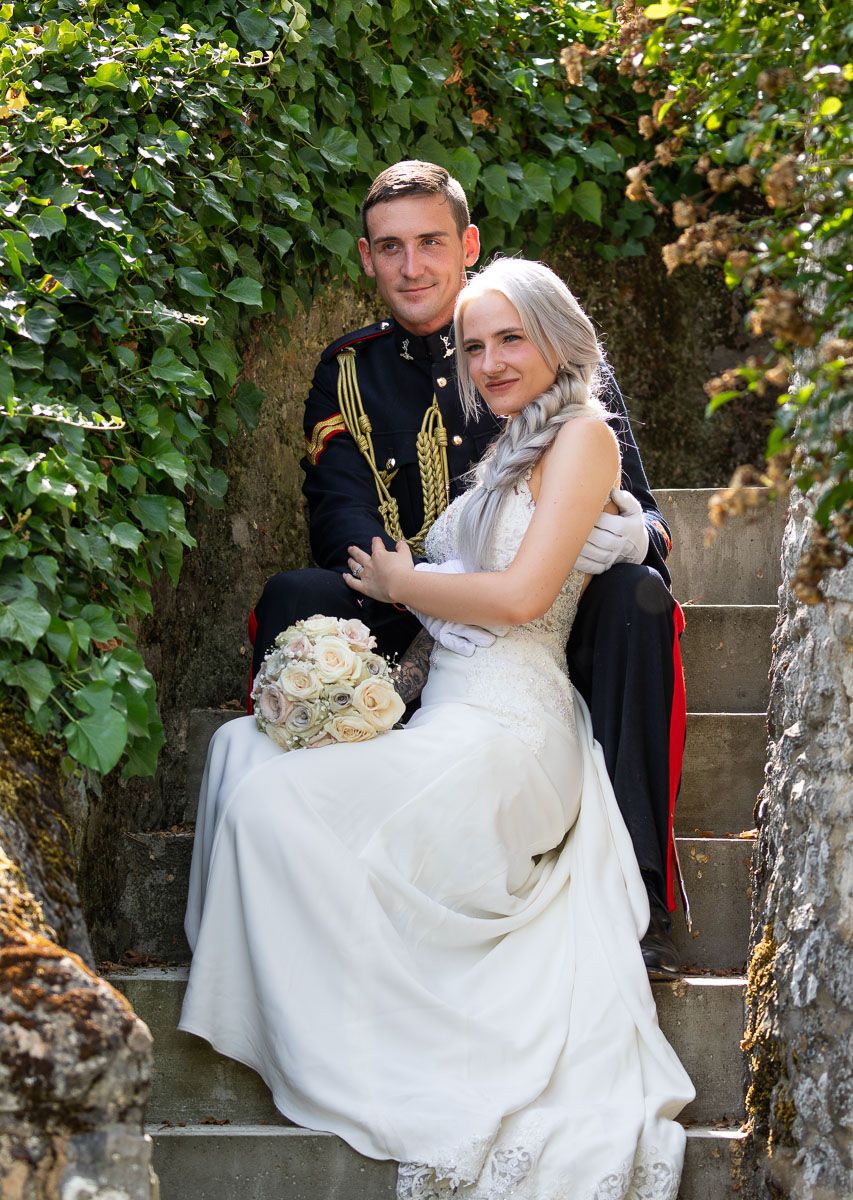 A couple in wedding attire poses together on stone steps of Allington Castle surrounded by lush green foliage in a romantic outdoor setting.