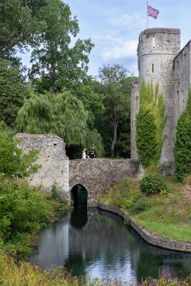 Wedding couple walk across historic bridge at   Allington Castle | stone castle walls and towers reflect in the tranquil moat.