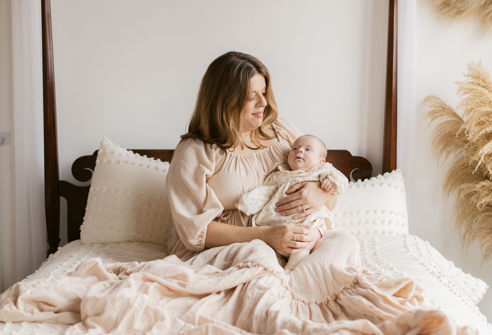A mother wearing a cream dress sits on a poster bed gazing at her newborn girl for newborn photos in Andover. Concord newborn photographer