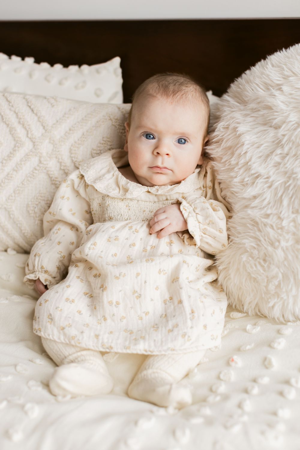 Newborn girl wearing a cream colored floral dress leans on pillows on a cream bedspread for newborn photos in North Andover, Massachusetts.