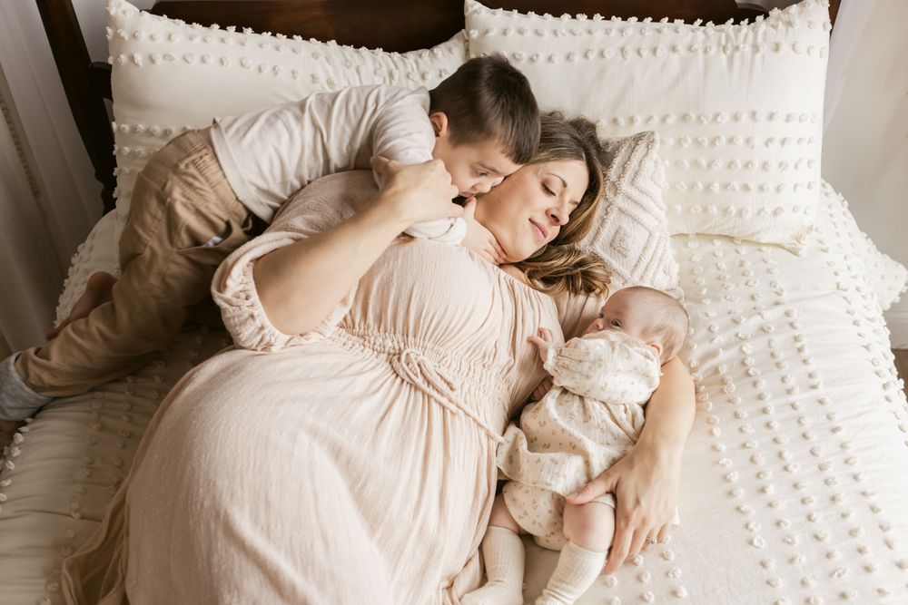 A preschool age boy embraces his mother as she lays on a bed with her newborn baby girl for family photos in North Andover, Massachusetts