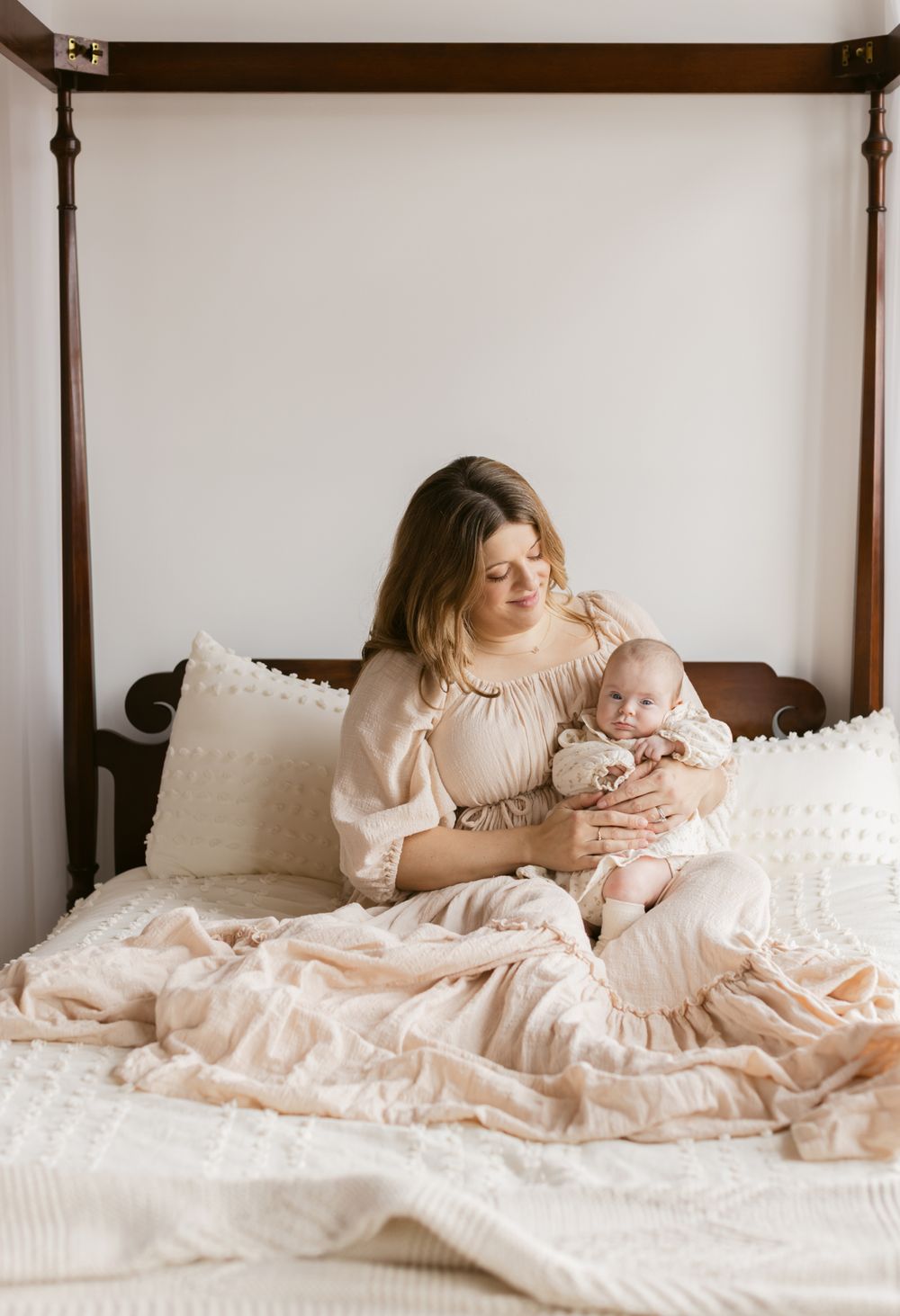 A mother wearing a cream colored dress holds her newborn girl on her arms sitting up on a poster bed for newborn photos in Andover, MA.