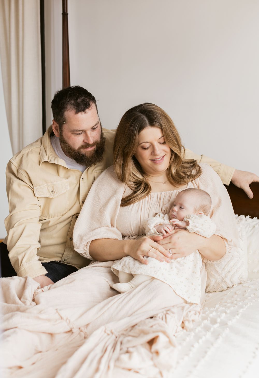Parents sit on a cream colored blanket poster bed holding their newborn daughter for newborn photos in North Andover, Massachusetts.