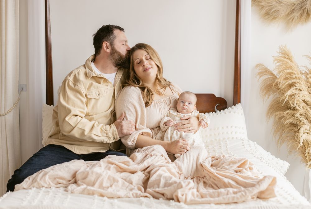 A husband kisses his wife's temple sitting together holding their newborn girl on a poster bed for newborn photos in North Andover, MA