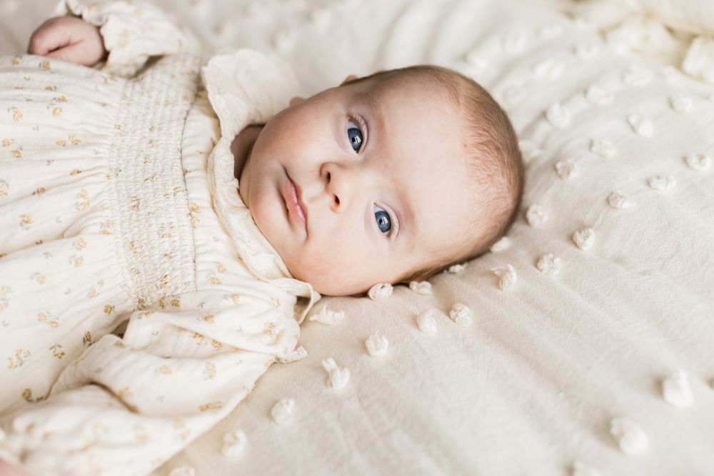 Newborn girl wears a cream floral dress gazes at camera for newborn photos laying on cream bedspread. Cohasset, Hampton newborn photographer