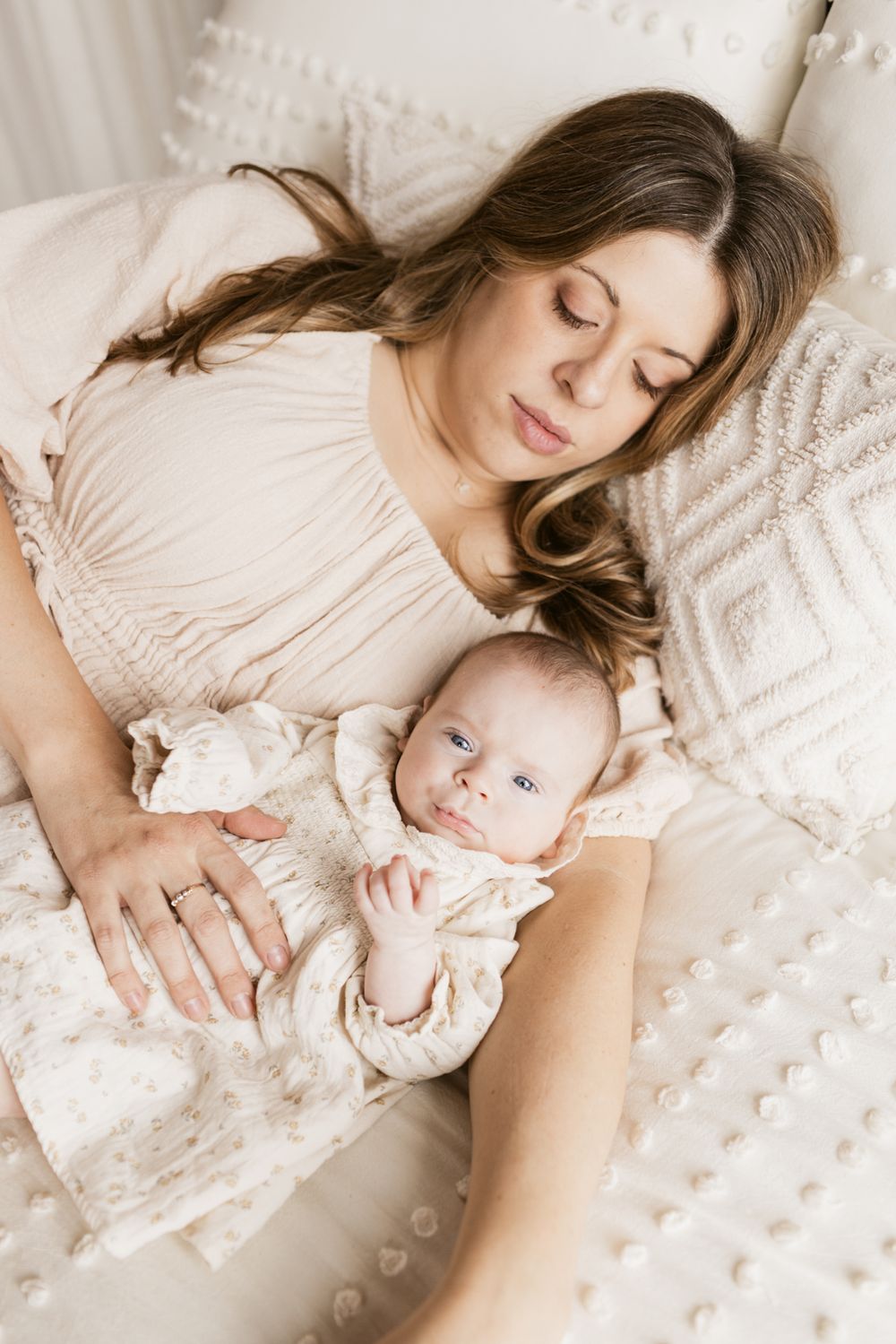 A mother with brown hair wearing a cream colored dress lays on a cream bedspread with her newborn girl snuggled next to her in North Andover