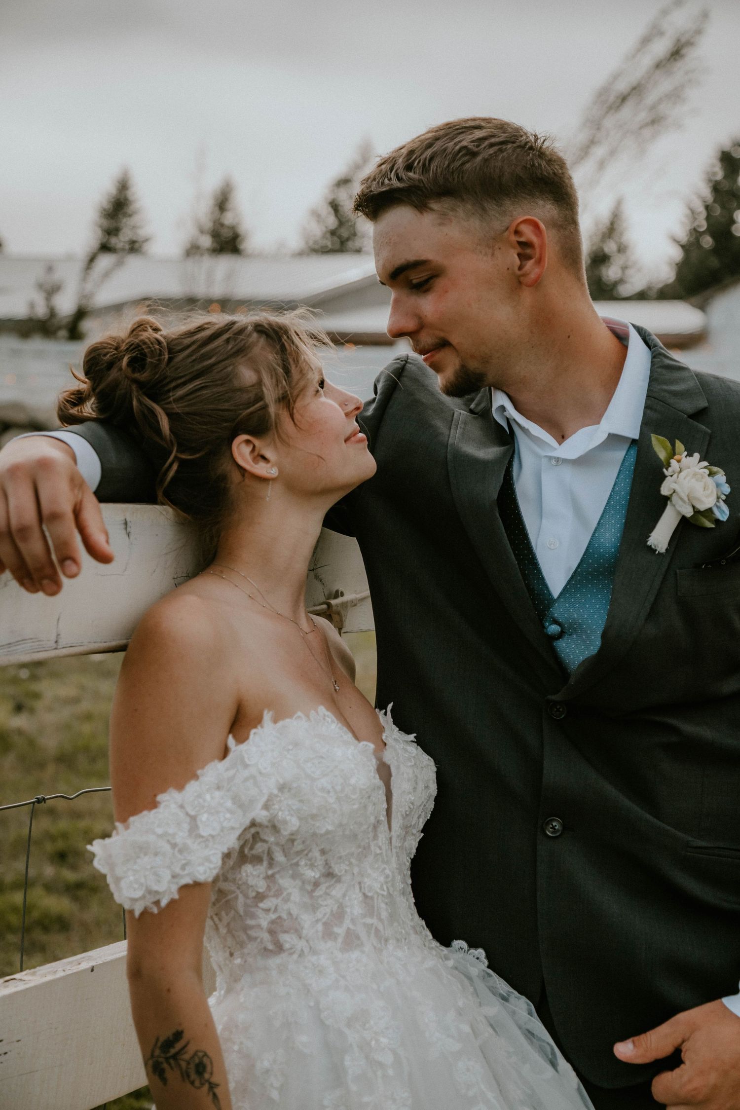 A romantic wedding couple shares an intimate moment outdoors with a rustic backdrop of evergreen trees.