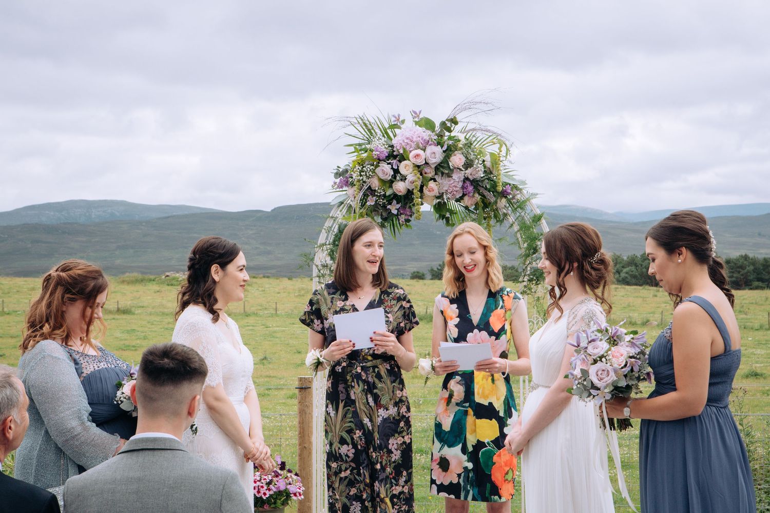 A wedding ceremony takes place outdoors with a floral arch and rolling green hills in the background.