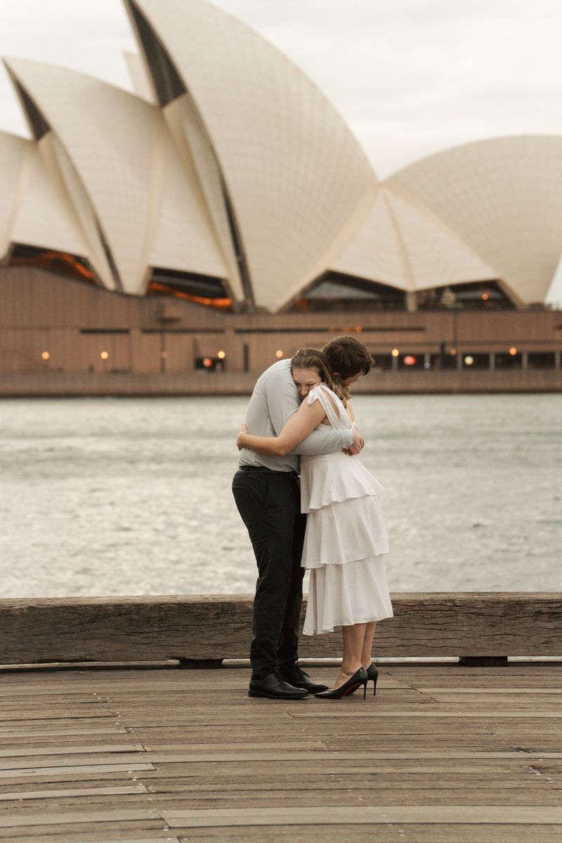 A couple embraces in front of the iconic Sydney Opera House on a cloudy day during a proposal photoshoot at the rocks