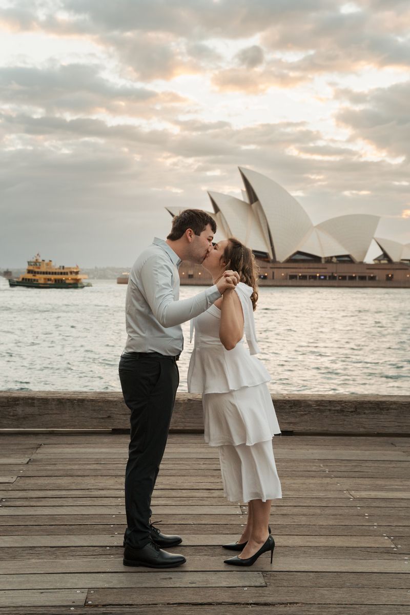 A romantic couple embraces on a wooden pier overlooking Sydney Harbour with the iconic Opera House in the background. Proposal Photoshoot