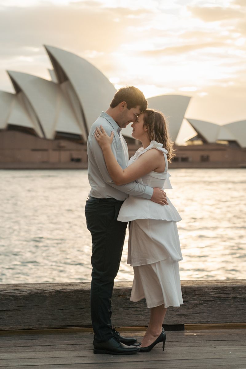 A couple shares a romantic kiss at sunrise with the Sydney Opera House silhouetted in the background during a proposal photoshoot
