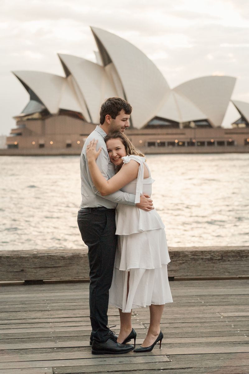 A romantic couple embraces on a deck overlooking the iconic Sydney Opera House during a cloudy day photoshoot at the rocks