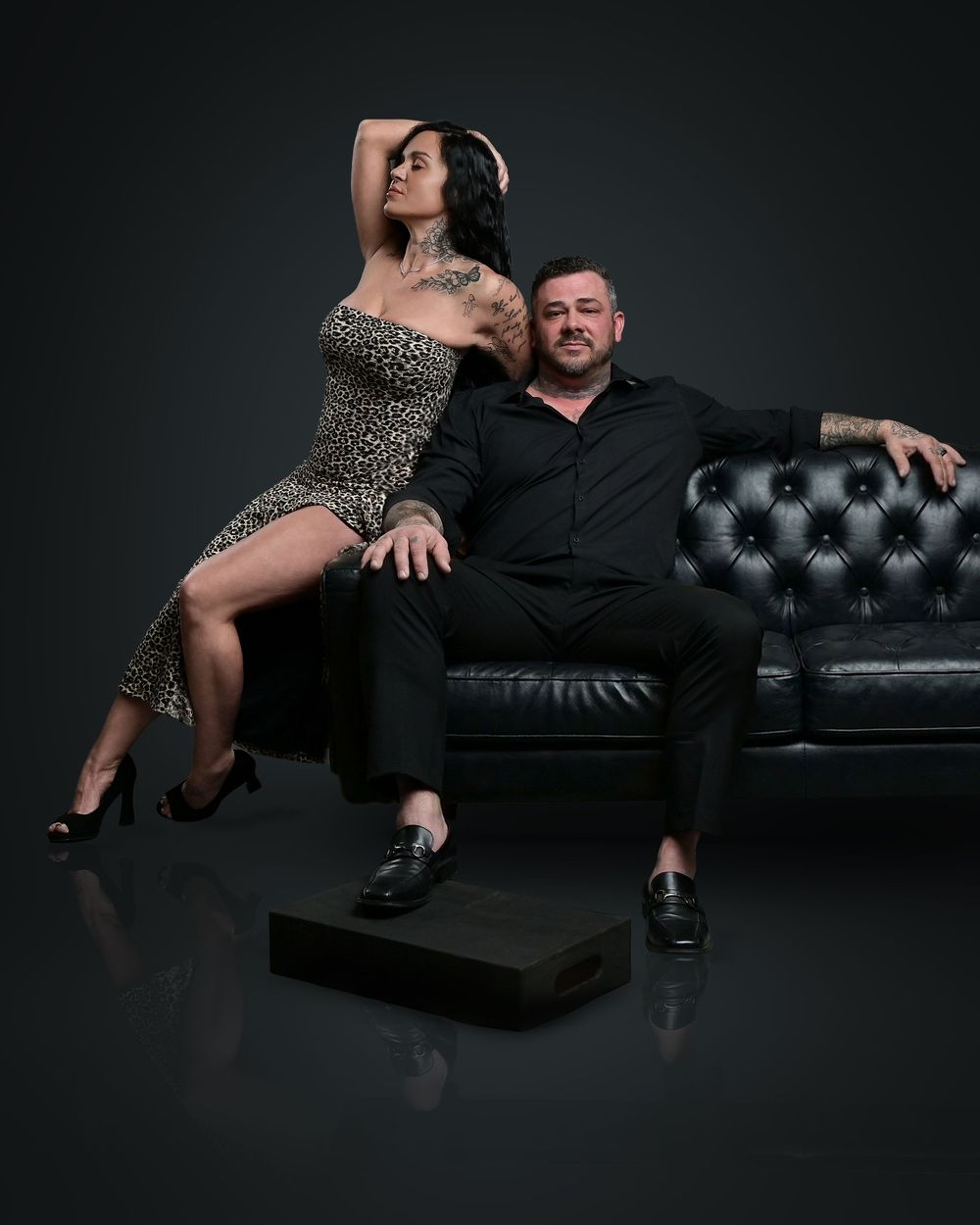 Glamorous couple posing on a black leather couch in formal attire.