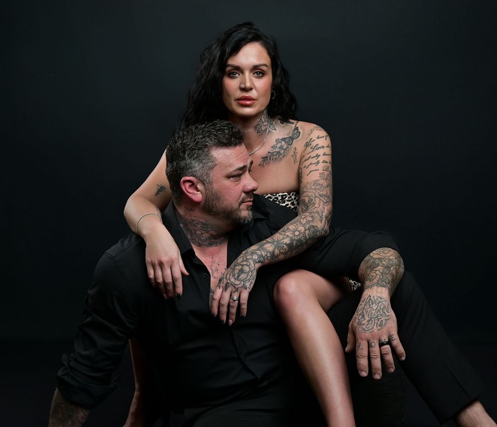 Artistic portrait of a tattooed couple in dark clothing against black background.