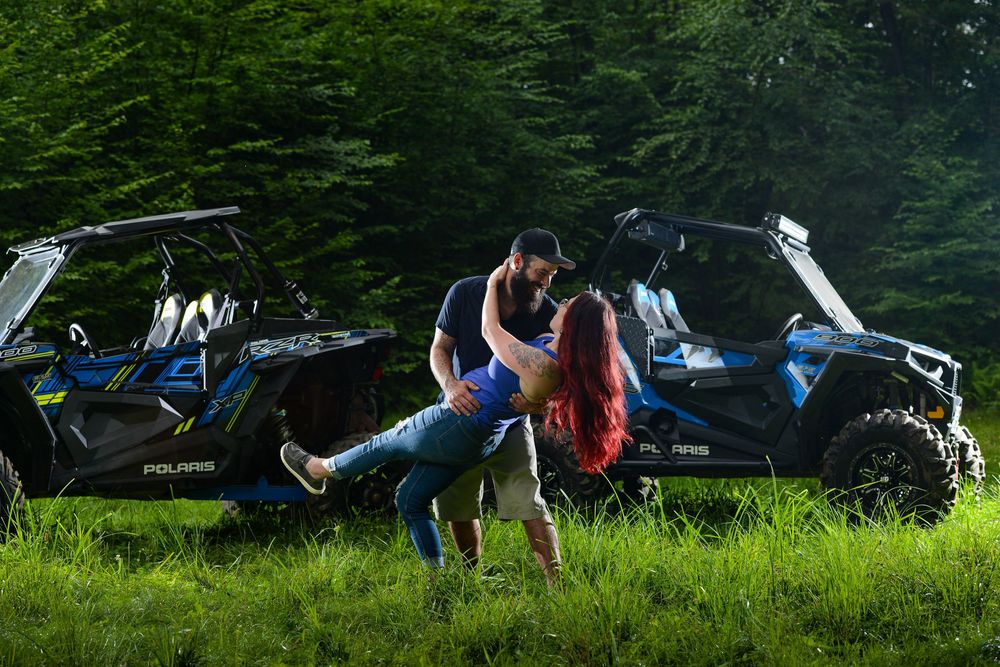 Couple in dramatic dip pose with off-road vehicles in background at sunset.