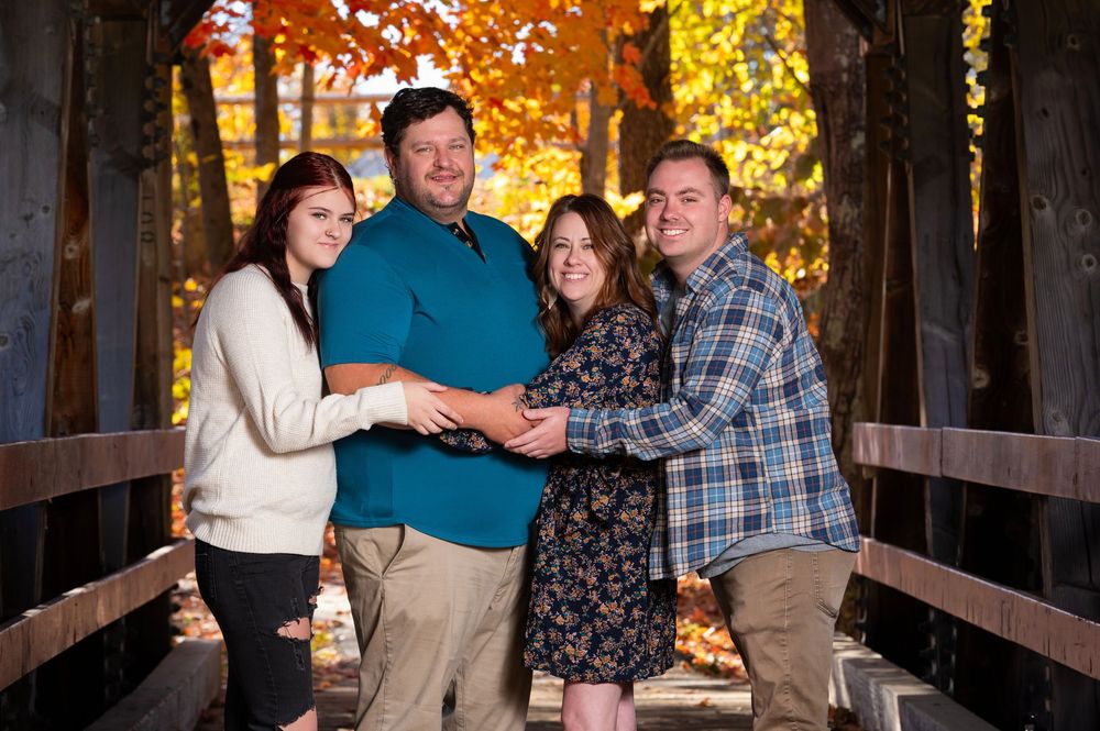 Family portrait of four people embracing on a wooden bridge with autumn foliage.
