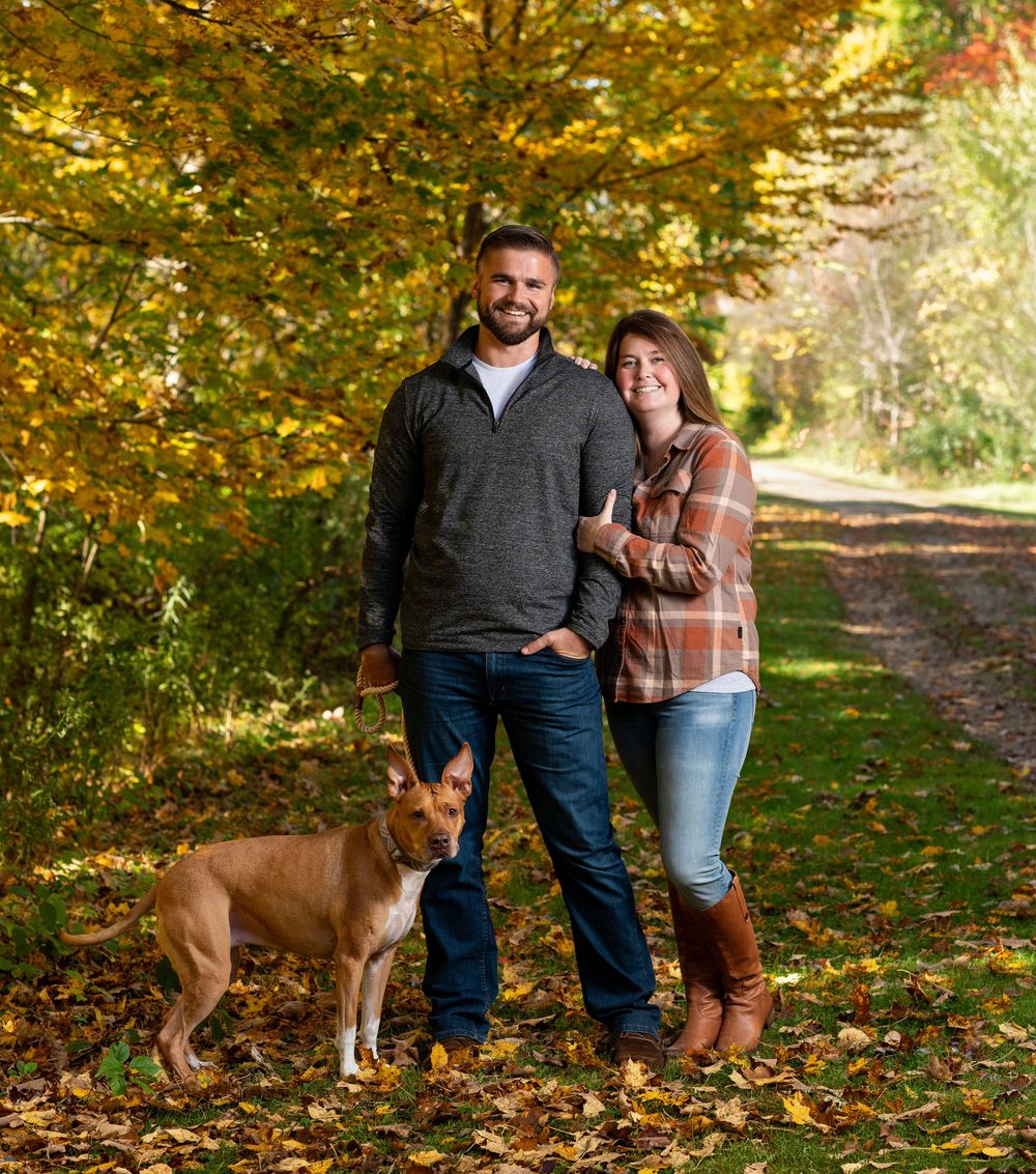 Couple and their brown dog posing together on a leaf-covered path during autumn.