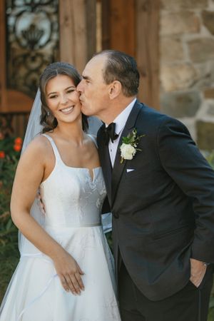 A tender moment between a bride in a white gown and a man in black suit at an outdoor wedding venue.