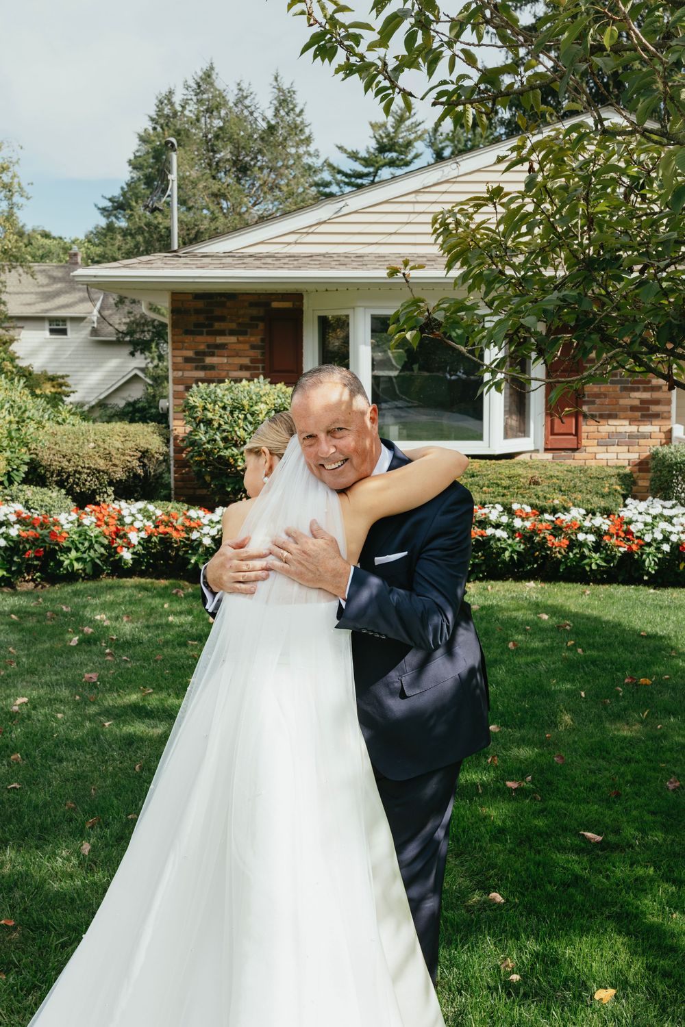 A couple shares an embrace in front of a ranch house with colorful flower gardens on a sunny day.