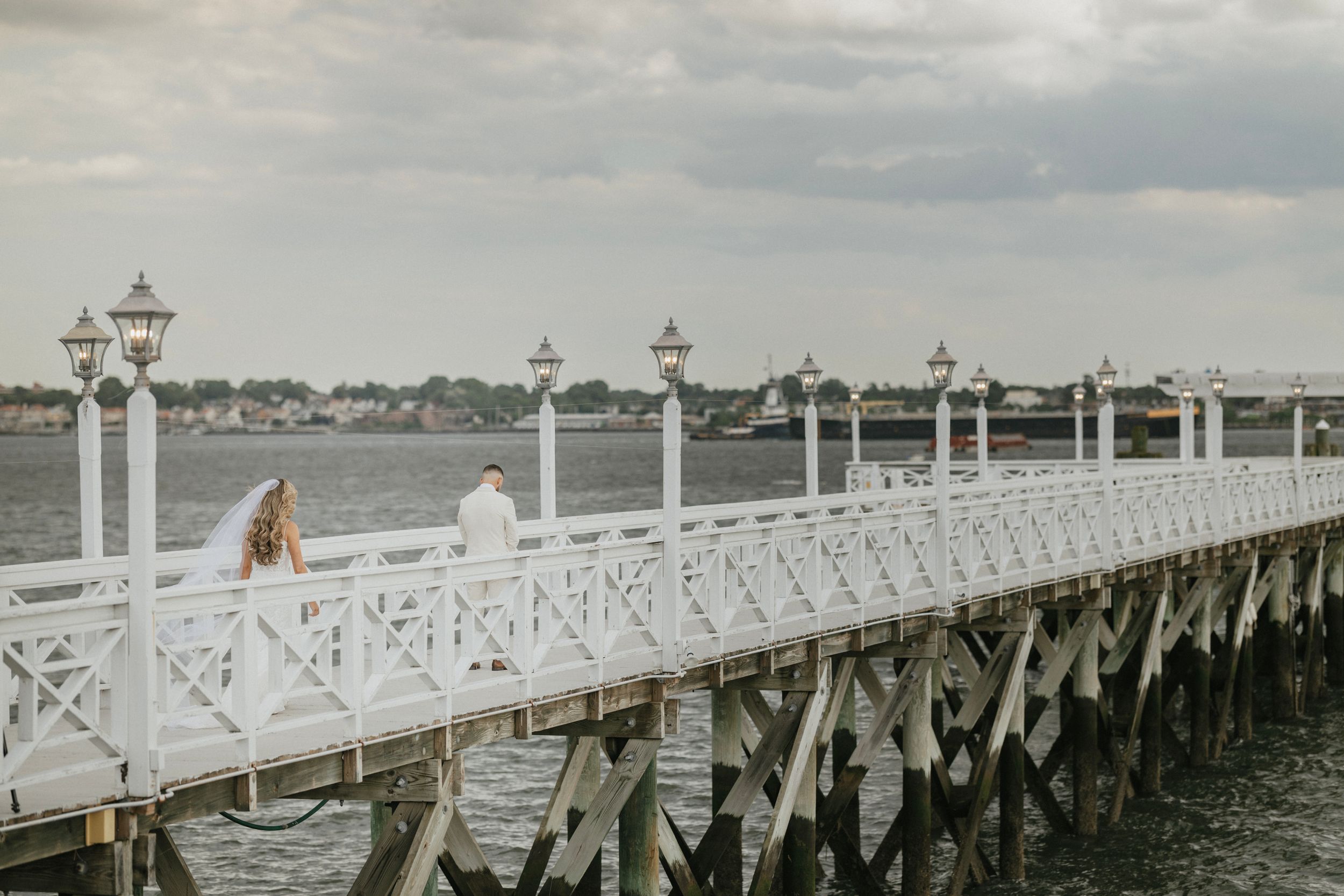 A white coastal pier with decorative lamp posts stretches over calm waters on a cloudy day.