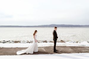 A couple stands apart on a snowy shoreline during a winter wedding photoshoot with a lake in the background.