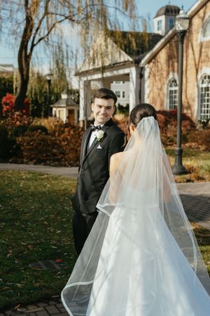 A couple shares their first look on their wedding day outside a historic building with willow trees in autumn.