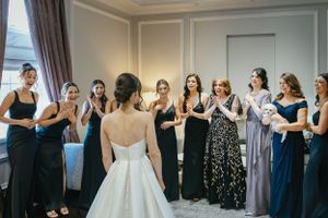 A group of bridesmaids in formal dresses gather around a bride in a white strapless wedding gown during preparations.