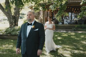 A groom in a dark suit stands in a garden setting while a bride in white waits behind him for a first look moment.