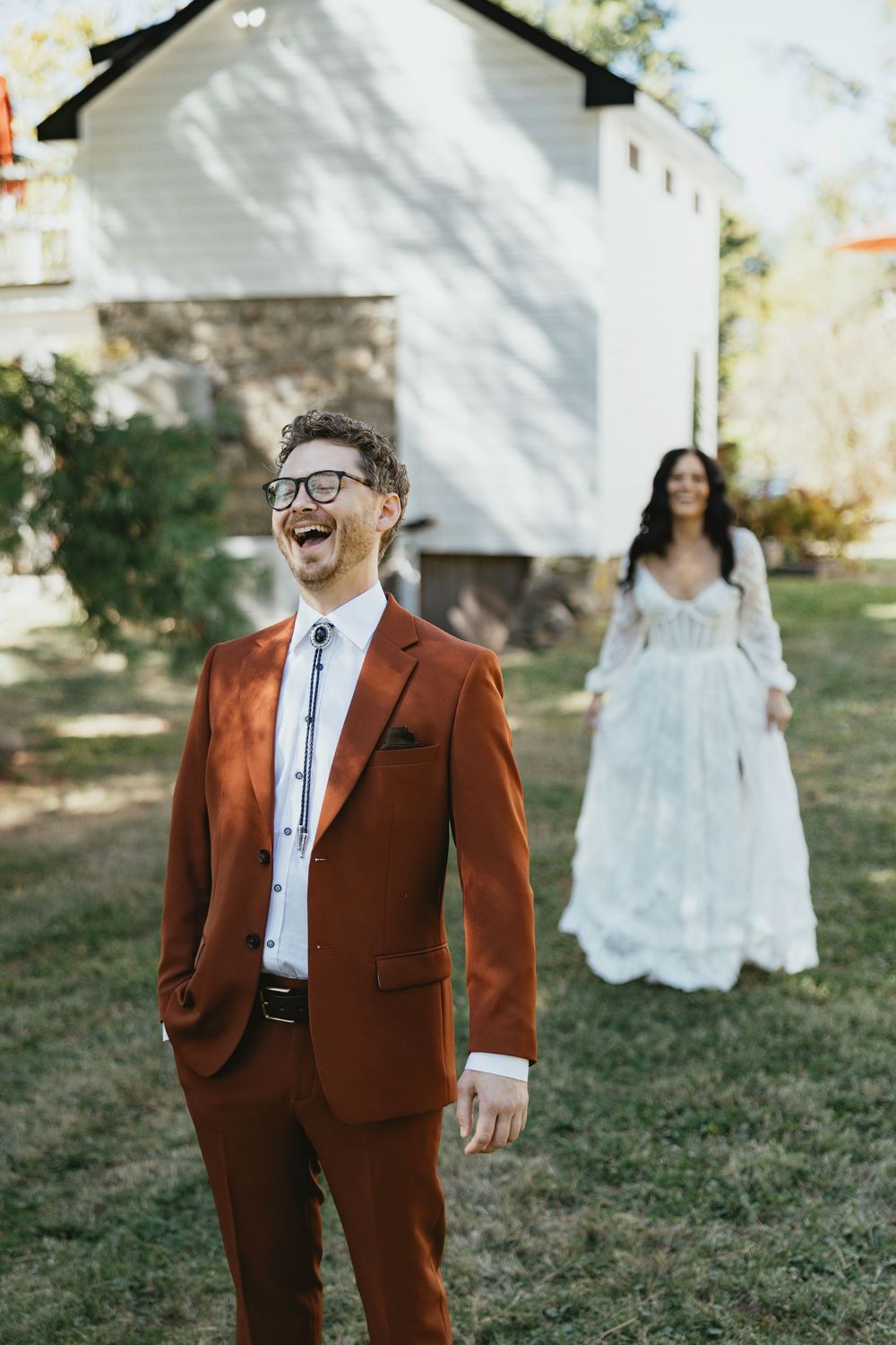 A joyful groom in a brown suit and glasses smiles during a first look moment with his bride in a garden setting.