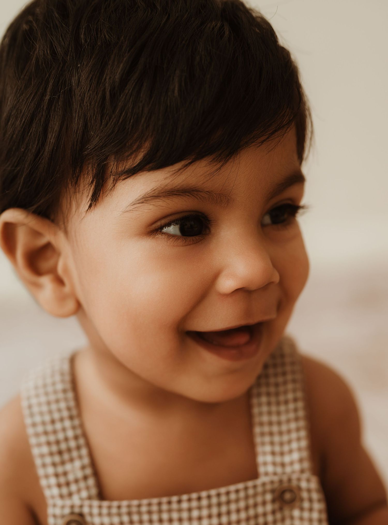 Baby in tan dress wearing small birthday hat sits on white rug with birthday balloons in warm-toned portrait setting.