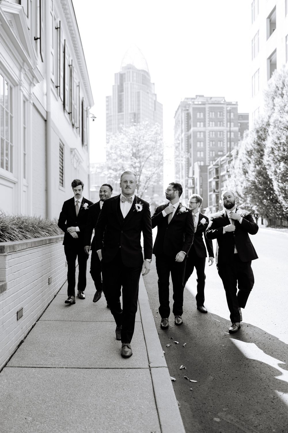 Groomsmen walking down urban sidewalk in formal black suits against city backdrop.