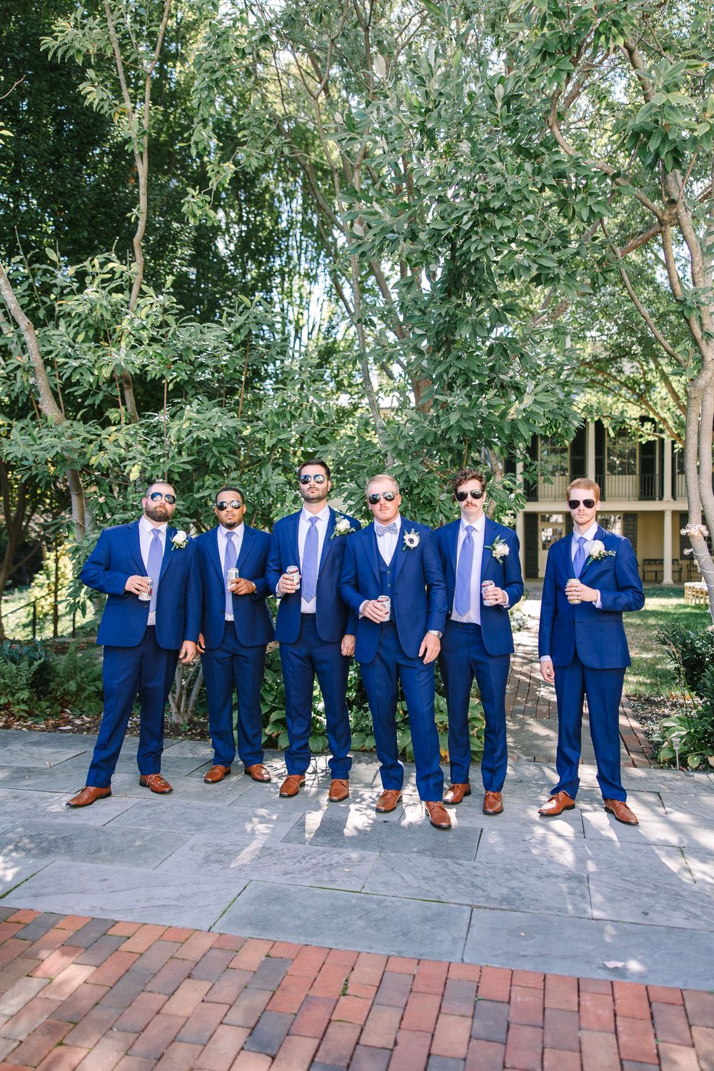 Groomsmen in matching navy suits and brown shoes pose together on a brick pathway surrounded by greenery.