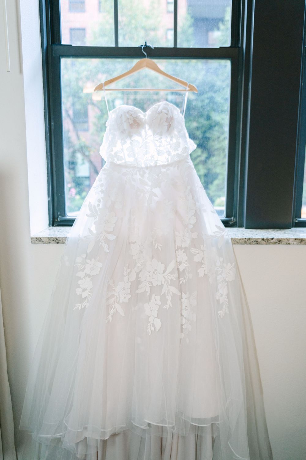 White wedding dresses hanging in front of bright windows in a bridal preparation room.
