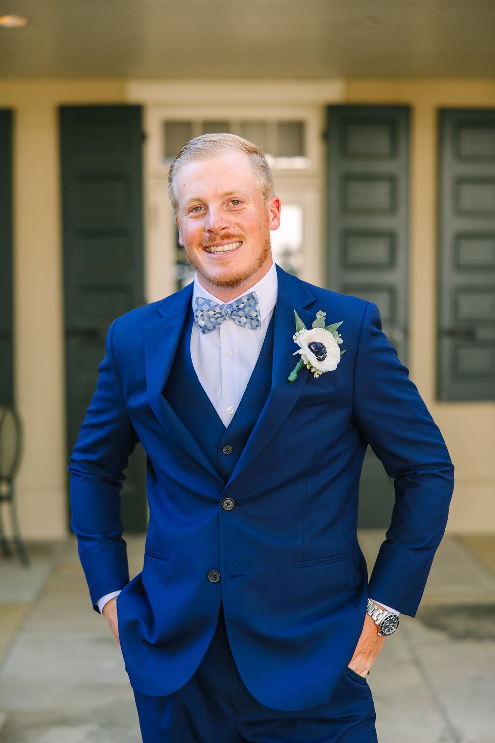 A person in a bright blue three-piece suit and white bowtie poses outdoors for wedding photos.