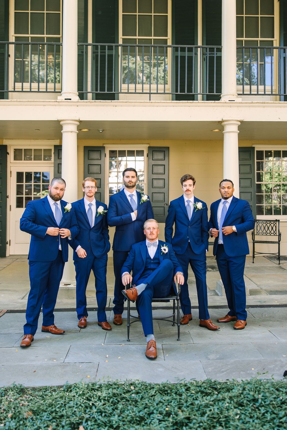 Groomsmen in navy blue suits and brown shoes pose casually on a porch with columns and windows.