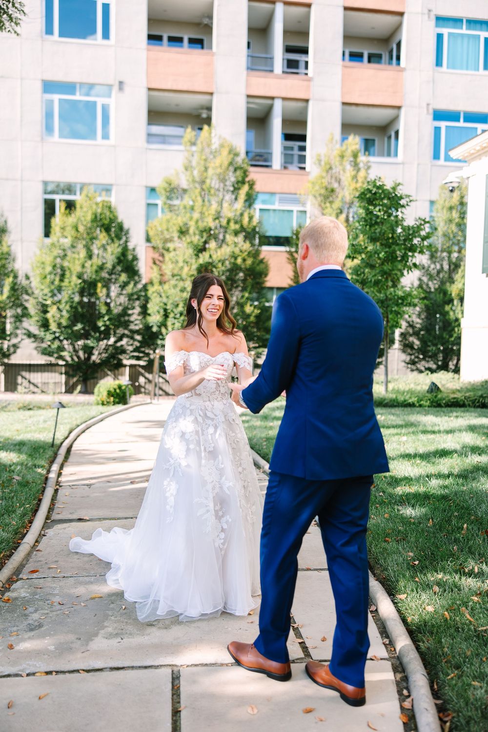 A bride in a lace off-shoulder gown and groom in navy suit share a moment on an outdoor walkway.